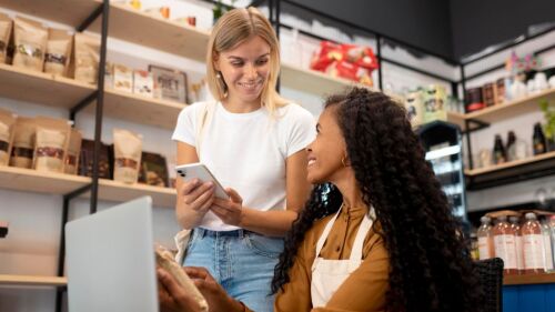 Mulher mostrando celular para outra mulher que está mexendo no computador; cena acontece em uma loja
