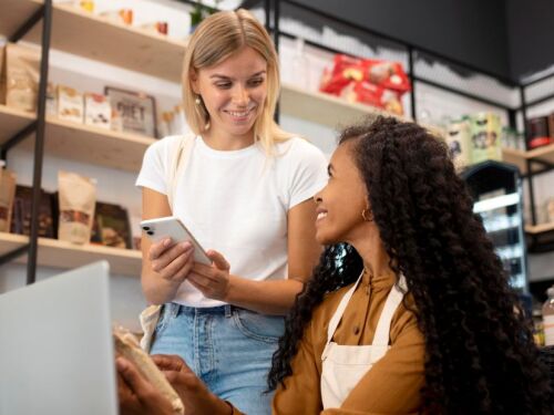 Mulher mostrando celular para outra mulher que está mexendo no computador; cena acontece em uma loja