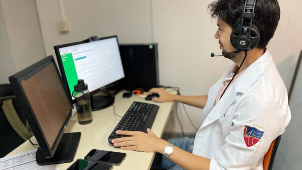 A imagem mostra um médico (um homem branco, de cabelo liso, na cor preta, e barba) sentado em frente à um computador, com um fone de ouvido na cabeça