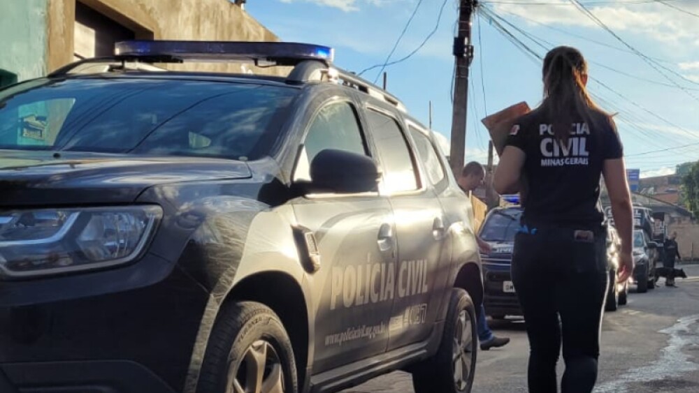 Mulher, de costas, com uniforme da polícia civil, ao lado de uma viatura da corporação